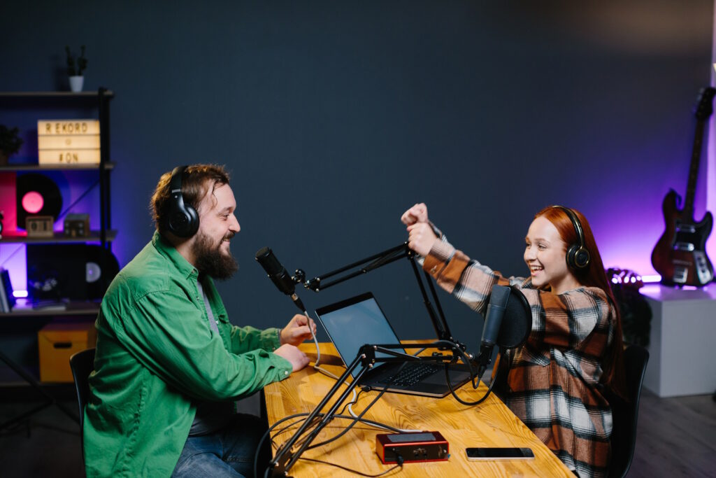 Two podcast hosts, a man in a green shirt and a woman in a plaid dress, share a fist bump in a studio adorned with musical instruments, reflecting the fun and excitement of bringing podcast characters to life.