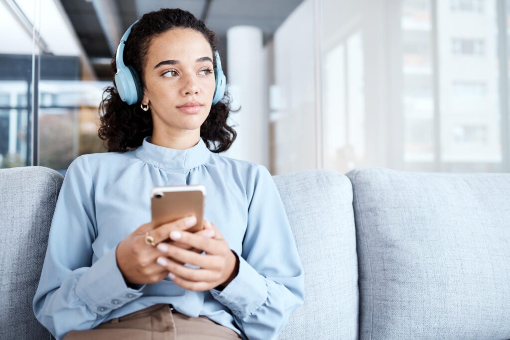 A thoughtful woman wearing headphones holds her smartphone, potentially pausing on a climactic moment as she experiences the thrill of building suspense in podcast .